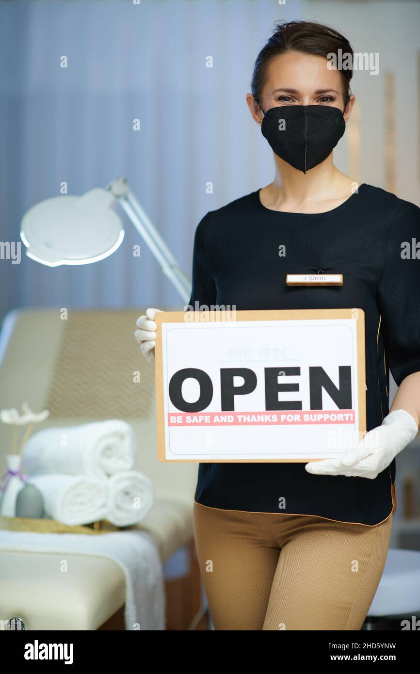Business during coronavirus pandemic. middle aged woman employee with ffp2 mask and open sign in modern beauty studio. Stock Photo