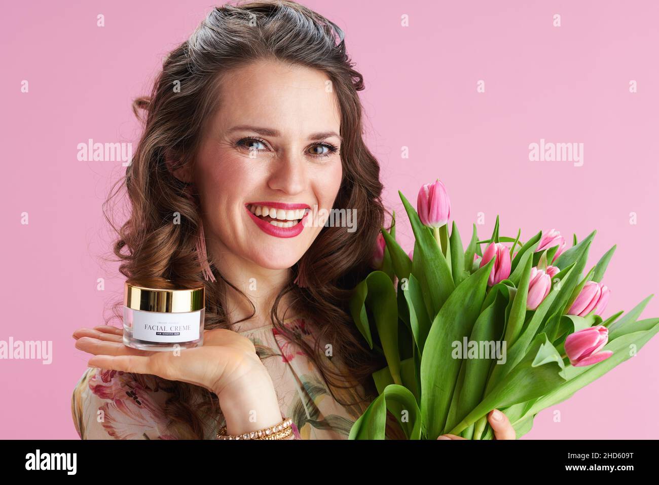 Portrait of smiling elegant female in floral dress with tulips bouquet and facial creme isolated on pink. Stock Photo