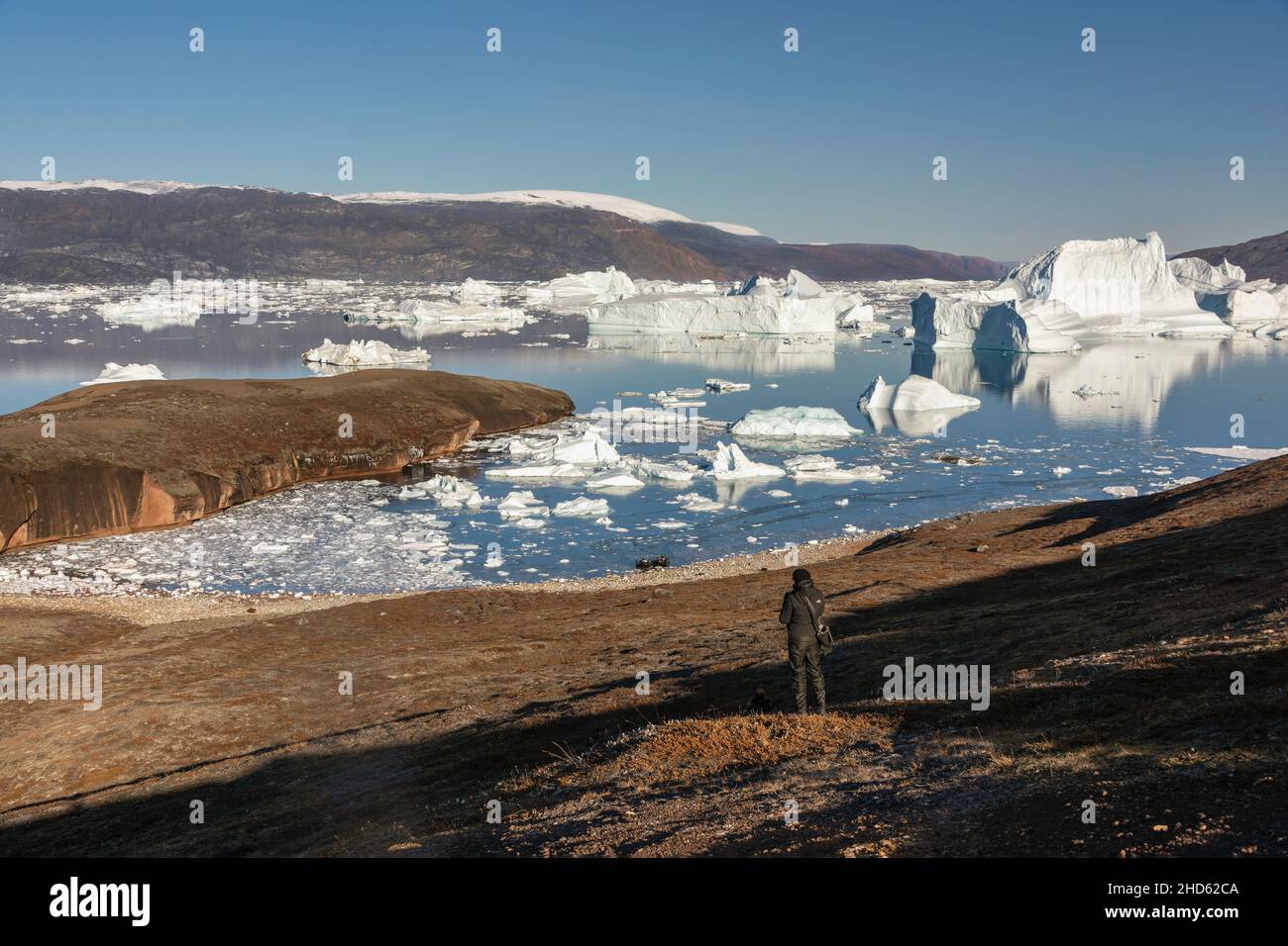 Photographer on Rode O looking down towards the Rodefjord beach with zodiacs and icebergs, Scoresby Sund, Greenland Stock Photo