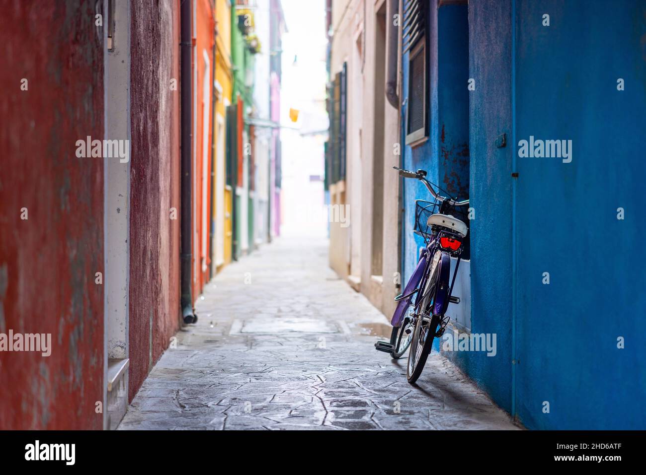 A purple bicycle is left leaning on a blue wall in a colorful backalley in Burano Stock Photo