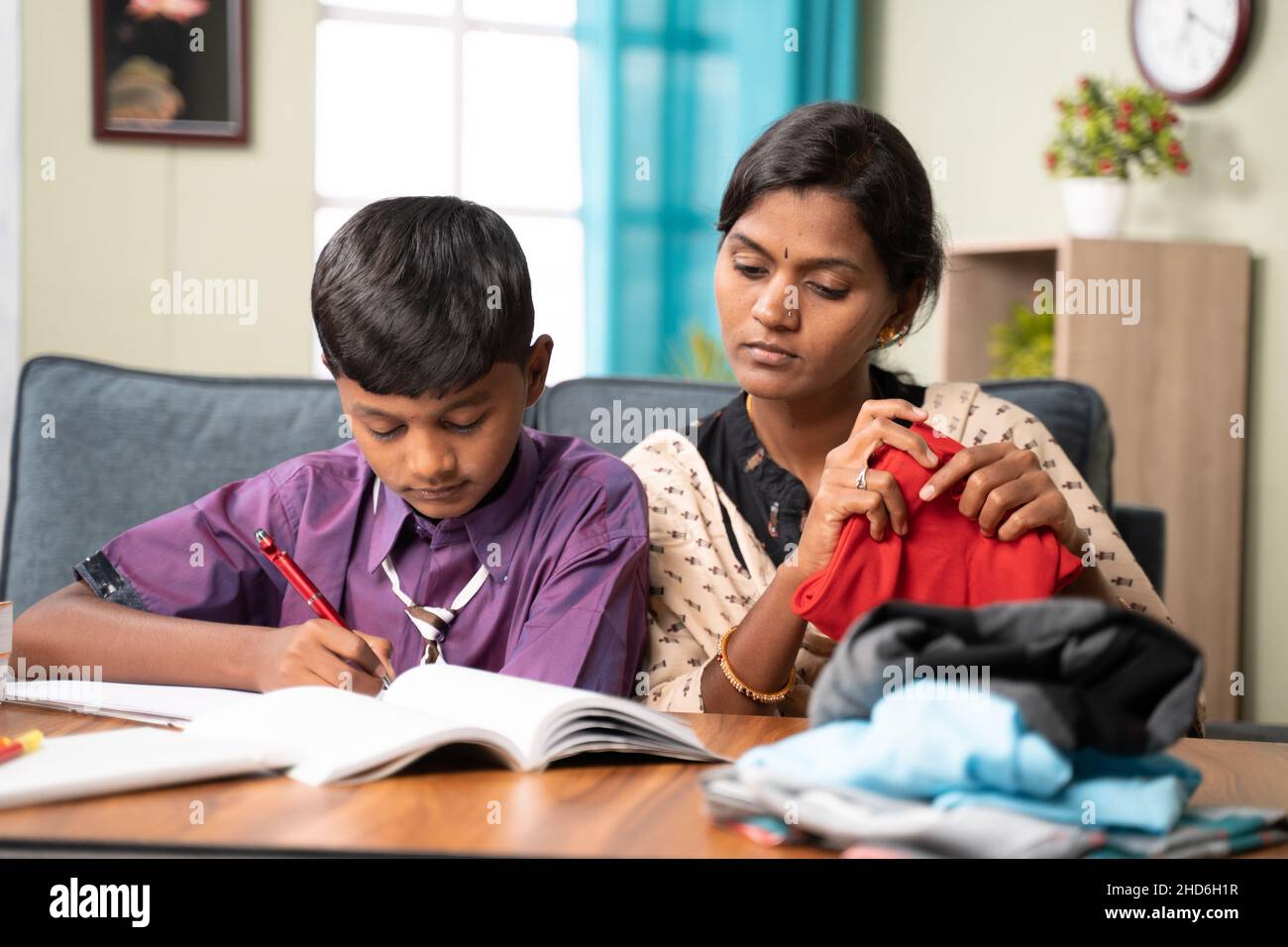 Focus on son, Mother helping her child to do homework while busy folding cloths at home - concept of education, learning, relationship and bonding Stock Photo