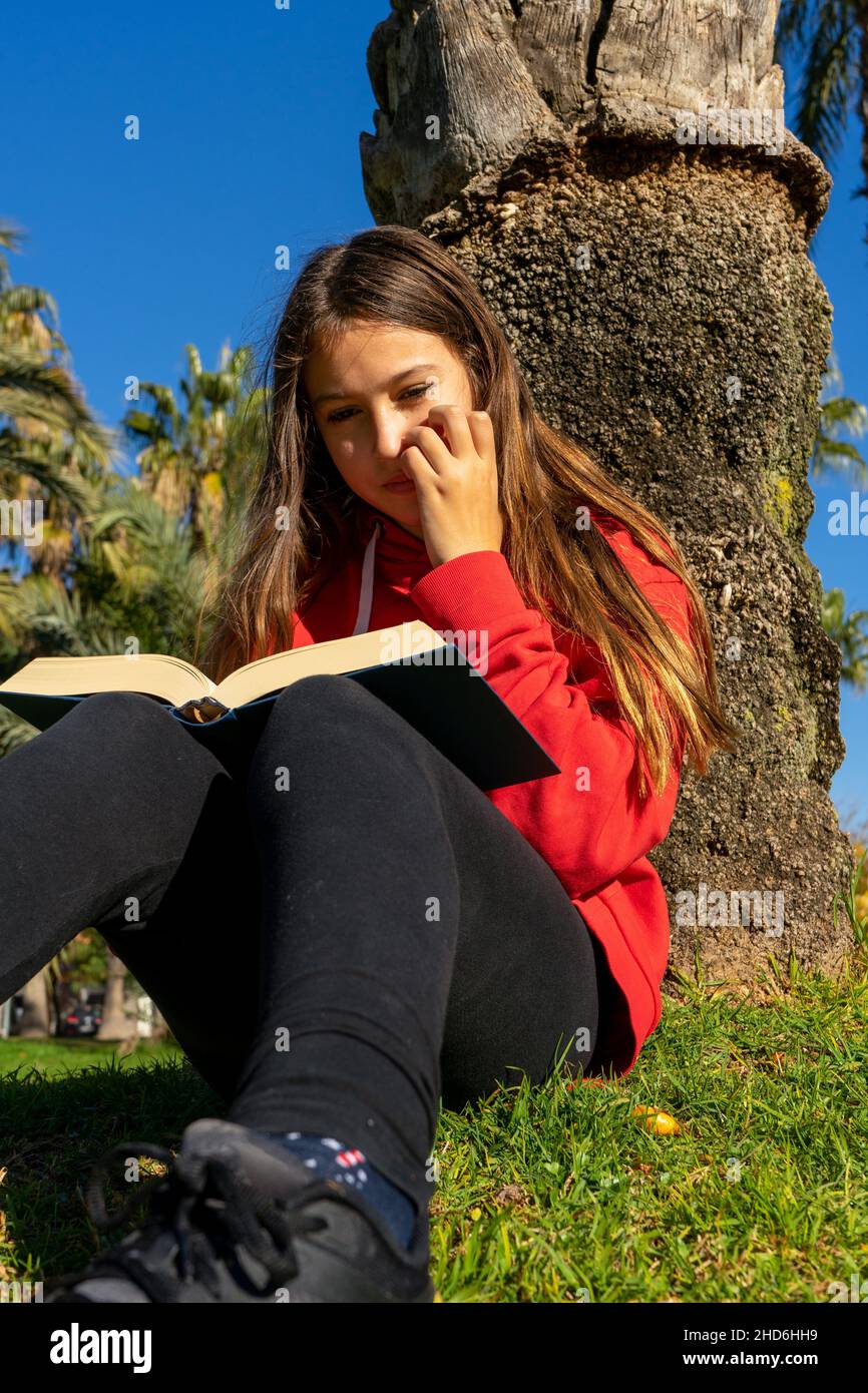 Pretty Spanish preteen girl reading a book on the green grass leaning against a tree in the park. Enjoy her hobby on a sunny winter day. Stock Photo