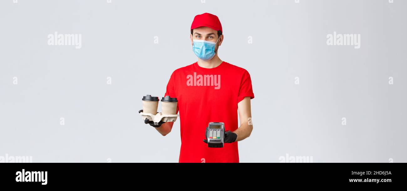 Takeaway, food and groceries delivery, covid-19 contactless orders concept. Pleasant courier in red uniform, gloves and face mask, holding coffee for Stock Photo