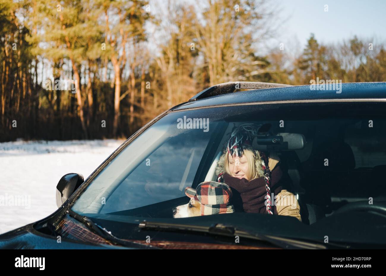 Happy woman with a dog wearing funny winter hats in the car by the forest. Winter outdoor scene with pet, lifestyle with dogs Stock Photo