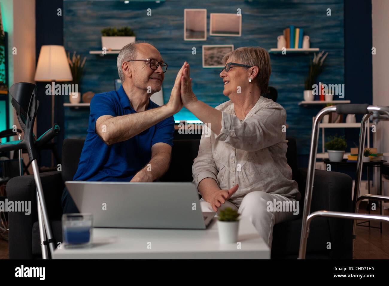 Caucasian old people highfive while sitting on sofa using laptop at home. Senior couple with technology and modern gadget enjoying retirement together indoors. Man and woman in living room Stock Photo