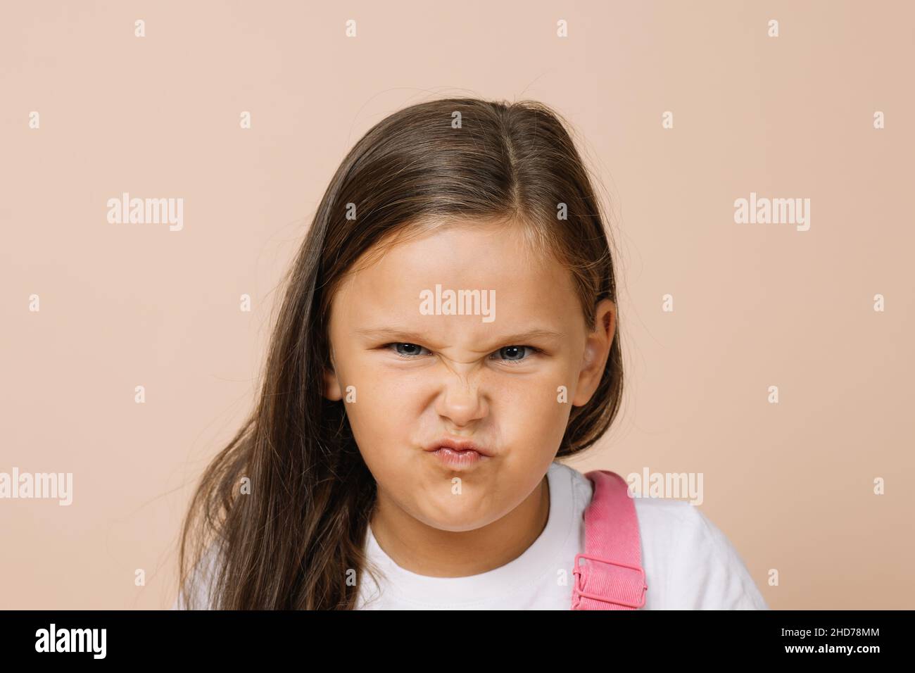 Portrait child with dissatisfied grimacing face and displeased angry eyes looking at camera wearing bright pink jumpsuit and white t-shirt on beige Stock Photo