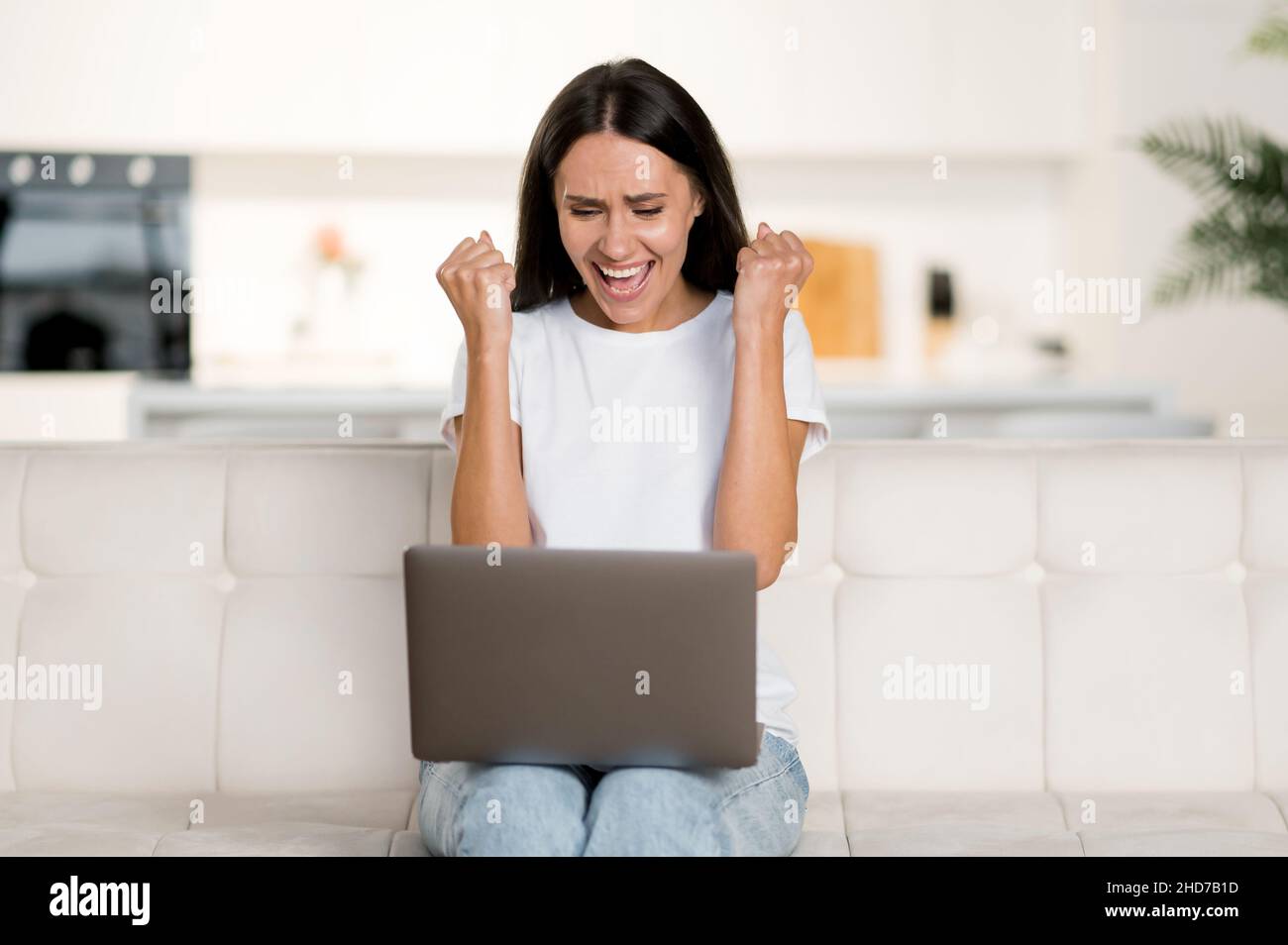 Happy young cute confident brunette caucasian woman, in casual clothes, sitting on sofa, using laptop, celebrating good profit, rejoice news or message, gesturing with fists, looking at screen, smile Stock Photo