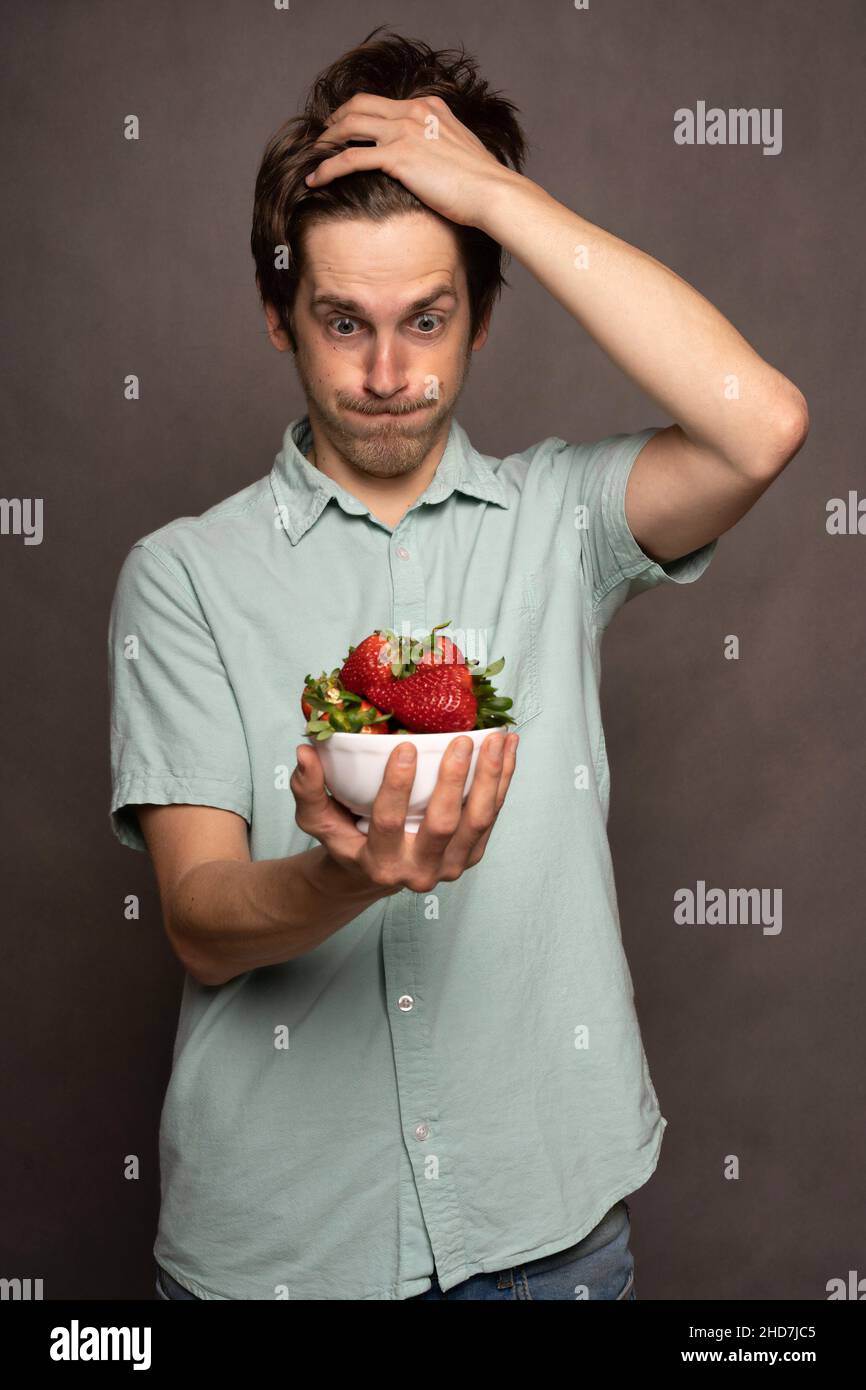 Young handsome tall slim white man with brown hair holding strawberries speechless in light blue shirt on grey background Stock Photo