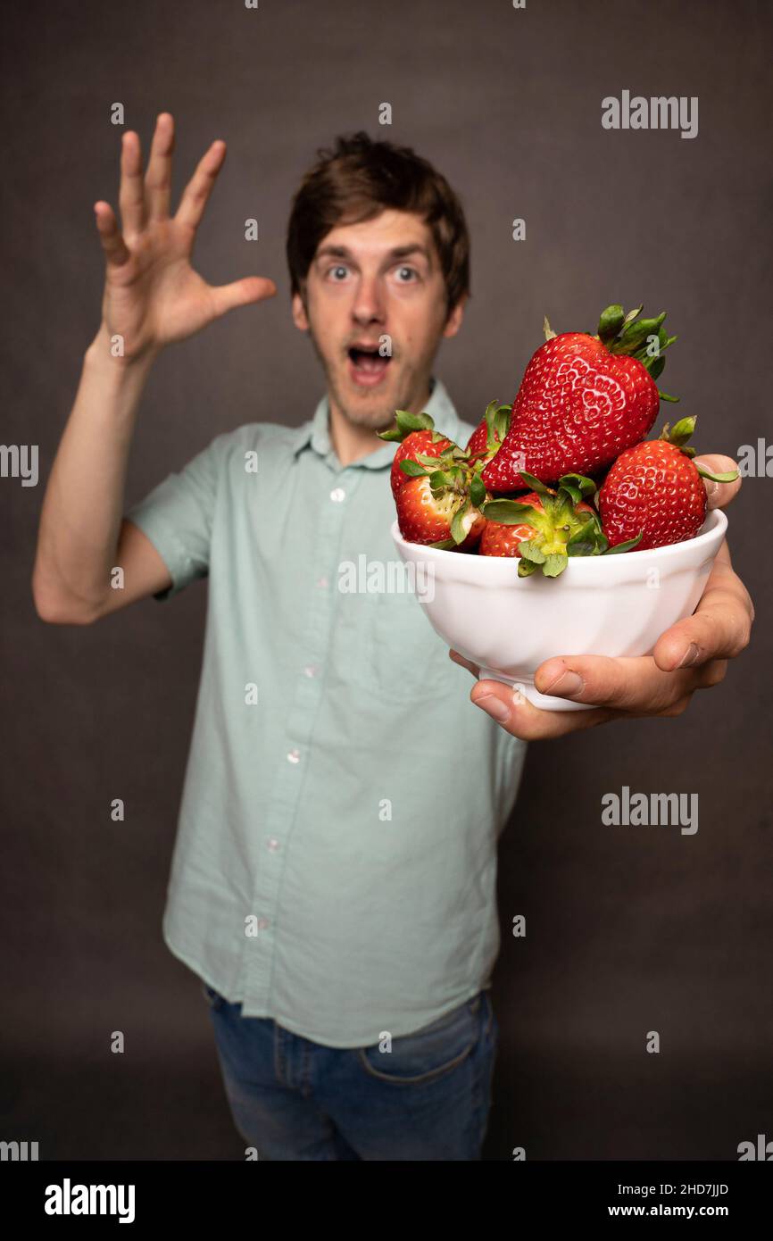 Young handsome tall slim white man with brown hair holding bunch of strawberries shocked in light blue shirt on grey background Stock Photo