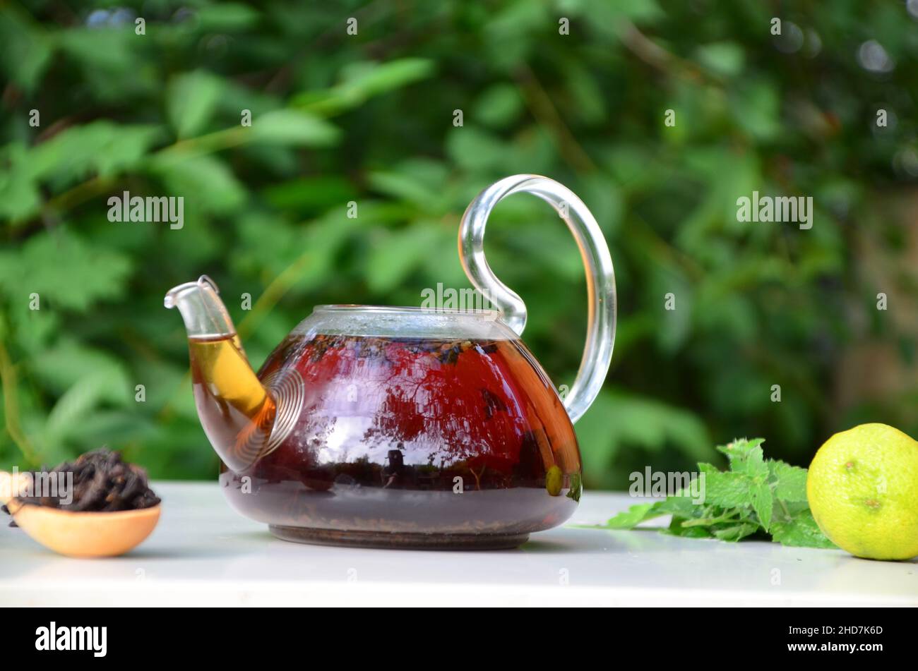 teapot full of tea on a background of green foliage on a wooden table, next to forest berries, raspberries and blueberries, lime or bergamot, mint. he Stock Photo