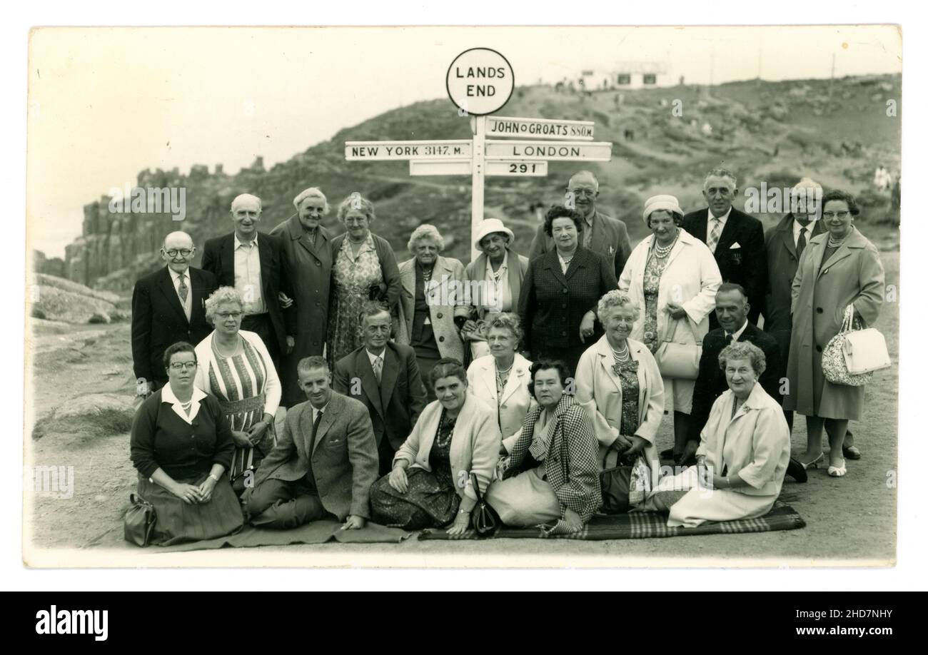 Original postcard of happy pensioners, men and women, wearing suits, carrying handbags, on an excursion in late Summer, probably coach tour group, to Land's End. They pose by the famous signpost, showing John O'Groats, London and New York mileage, printed on reverse Richards Bros. 3 Sept 1962, Cornwall, U.K. Stock Photo