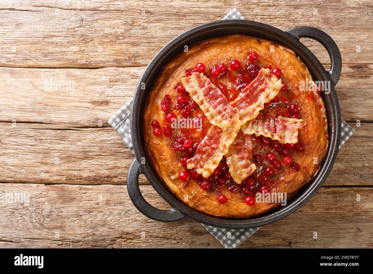 Aggakaga is a traditional South Swedish thick pancake with fried bacon and lingonberries closeup in the pan on the wooden table. Horizontal top view f Stock Photo