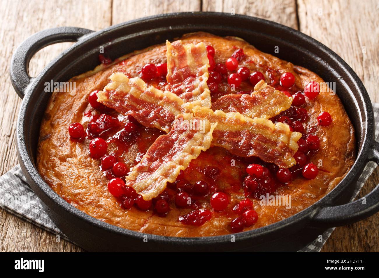 Aggakaga is a traditional South Swedish thick pancake with fried bacon and lingonberries closeup in the pan on the wooden table. Horizontal Stock Photo