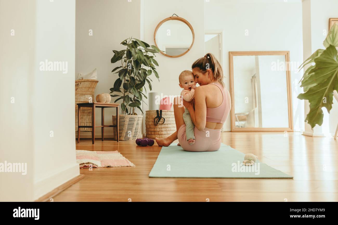Affectionate mom working out with her baby at home. Healthy mom holding her baby while sitting on an exercise mat. New mom bonding with her baby durin Stock Photo