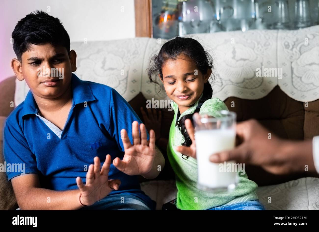 Indian teenager sibling kids saying no or refusing for drinking milk to parents while watching television at home - concept of unhealthy food habits Stock Photo