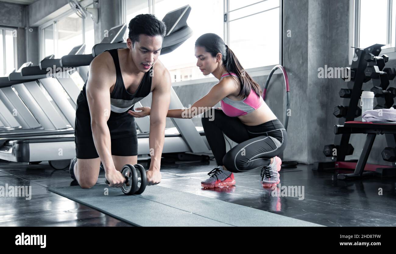 Handsome asian workout by ab wheel on yoga mat exercise with woman are trainers at gym. Concept of exercise in gym. Stock Photo