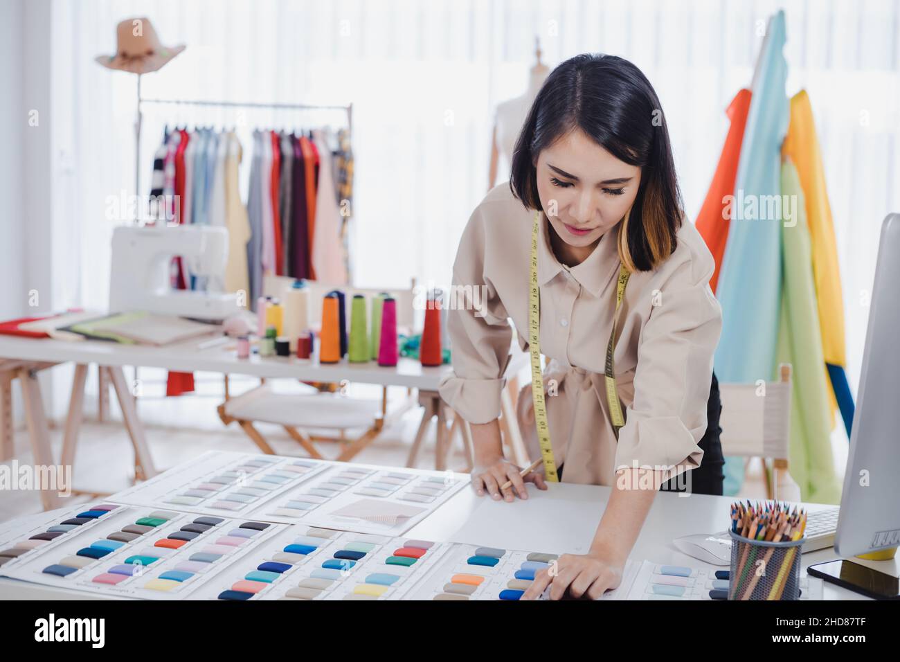 Asian woman designer are thinking and designing clothes for customers order items at the designer desk in the studio. Clothes designers are working in Stock Photo
