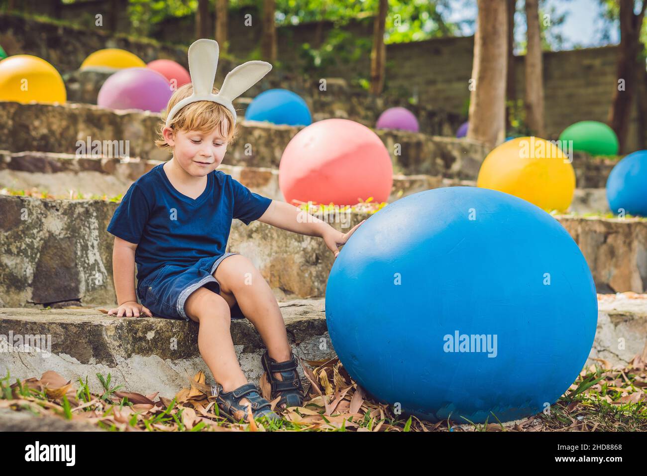 Cute little kid boy with bunny ears having fun with traditional Easter eggs hunt, outdoors. Celebrating Easter holiday. Toddler finding, colorful eggs Stock Photo