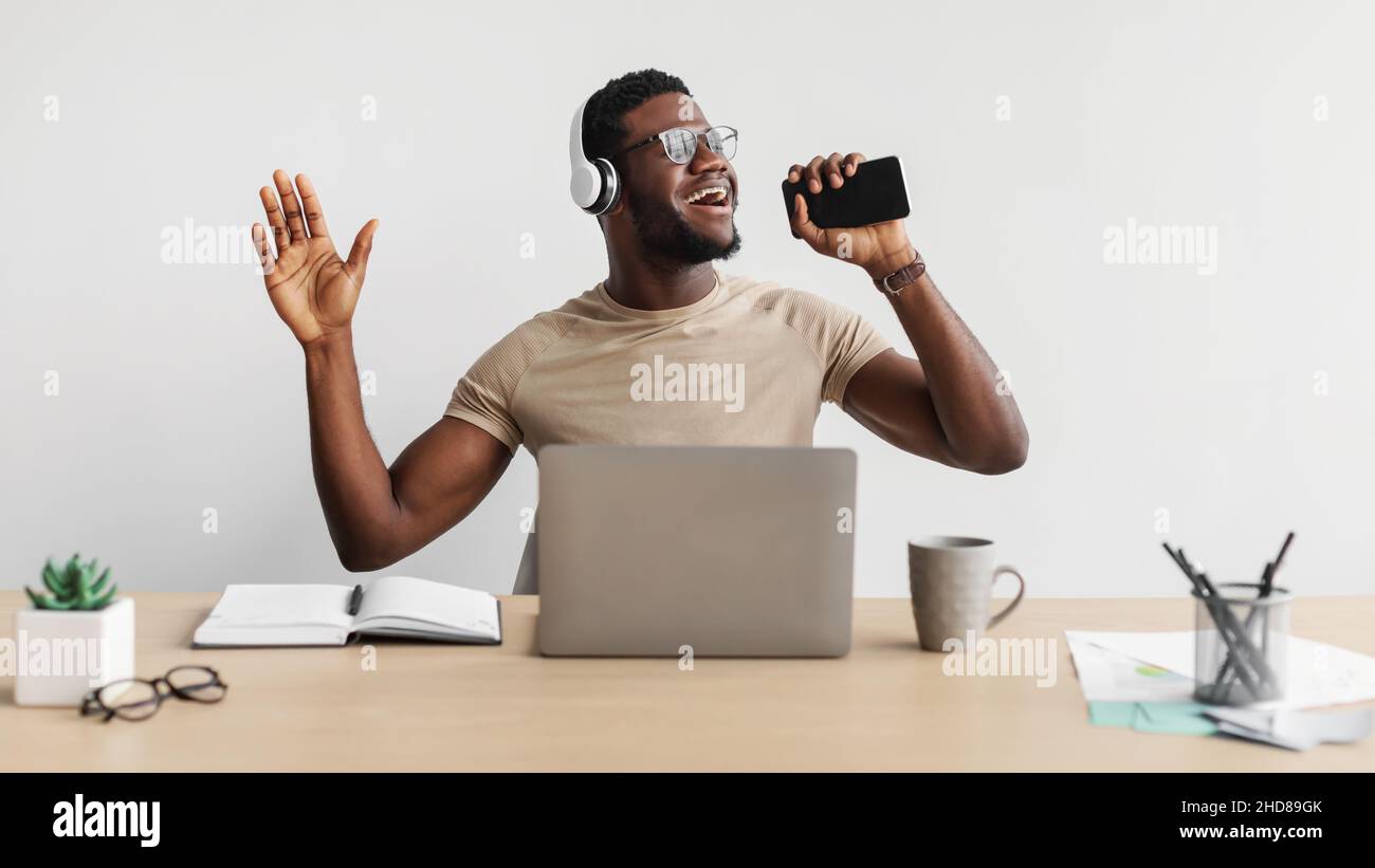 Cheerful young black man listening to music and singing into smartphone while working at desk on white background Stock Photo