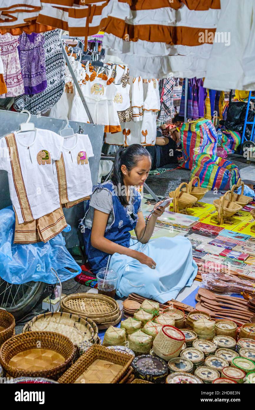 Pretty young girl stallholder in a souvenir stall looking at her mobile phone in the Walking street night market, central Luang Prabang, northern Laos Stock Photo