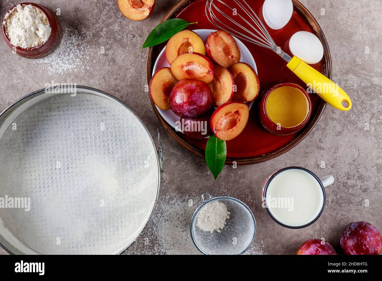 Baking process ingredients eggs, whisk, plum and baking pan on kitchen surface. Stock Photo