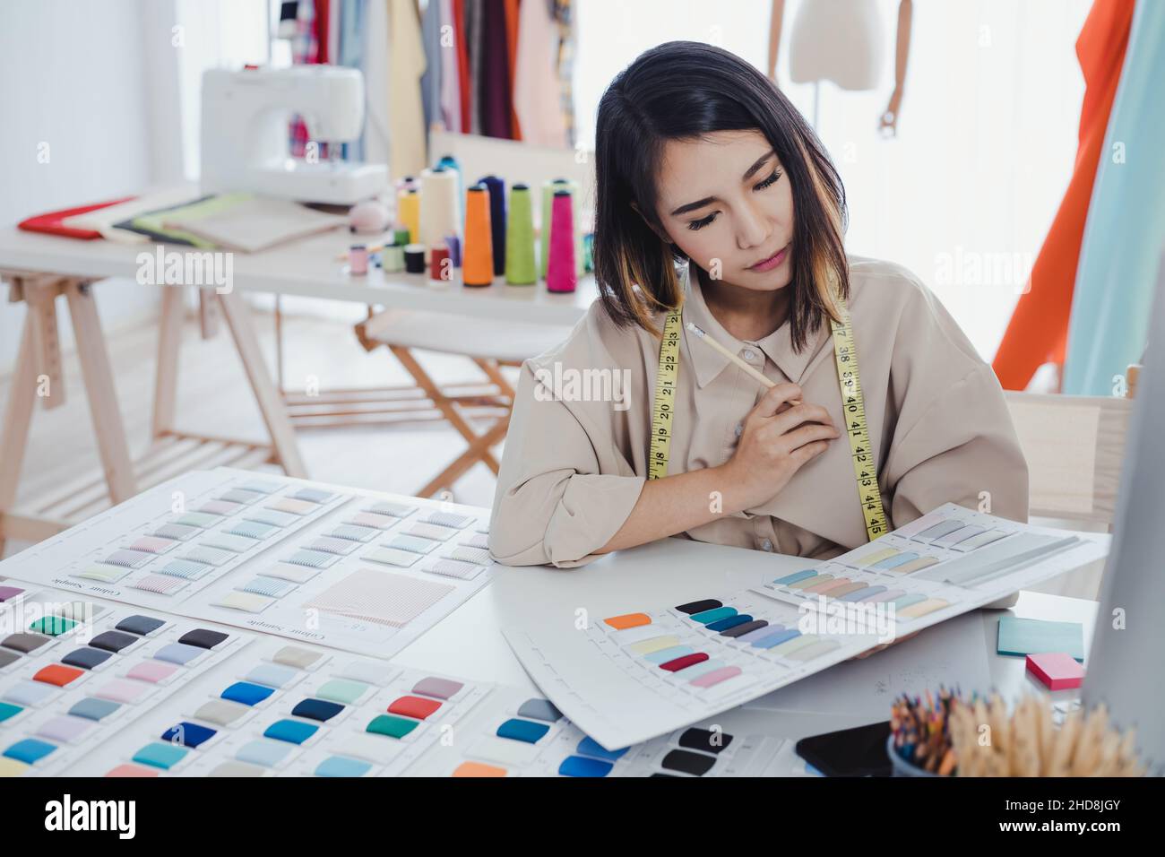 Asian woman designer are thinking and designing clothes for customers order items at the designer desk in the studio. Clothes designers are working in Stock Photo