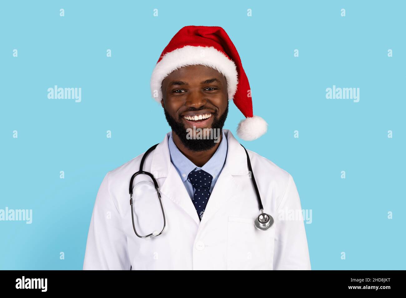 Handsome black doctor in uniform and Santa hat posing over blue background Stock Photo
