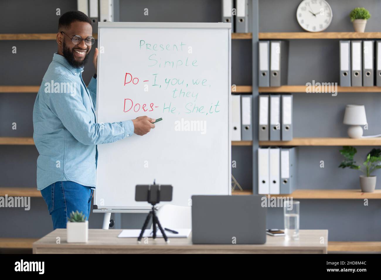 Happy millennial attractive black man tutor points to English rules on blackboard, looks at phone Stock Photo