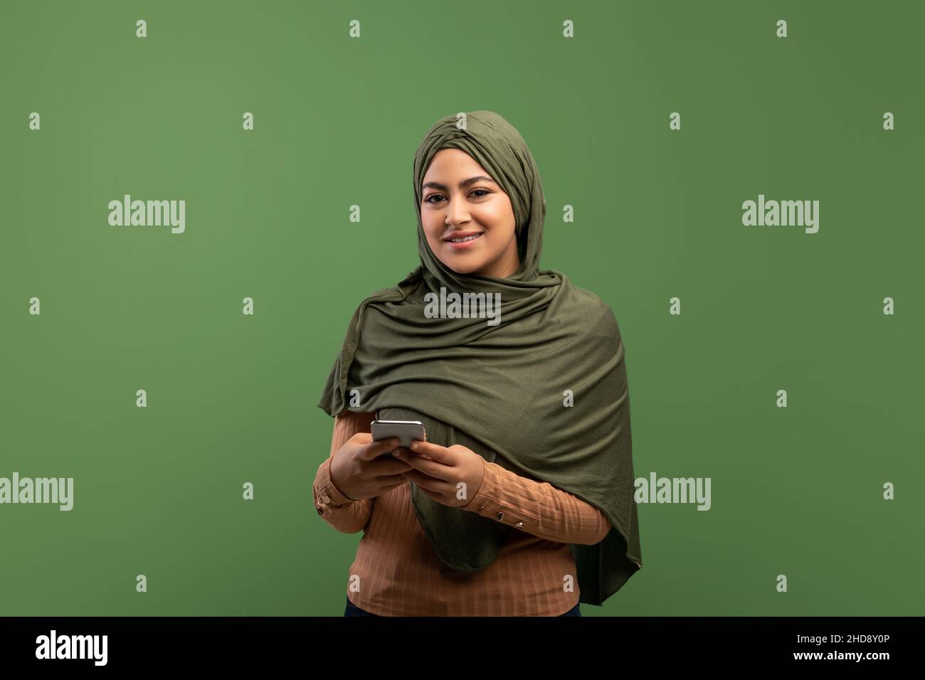 Cheerful muslim lady in hijab holding smartphone and smiling at camera ...