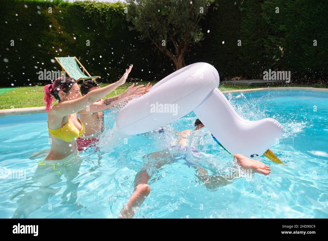 Two friends throwing man from a rainbow unicorn float into the pool. Stock Photo