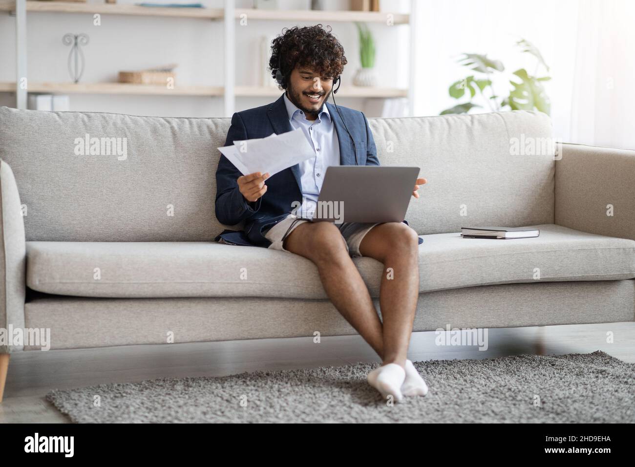 Attractive indian guy sitting on couch, using laptop and headset Stock Photo