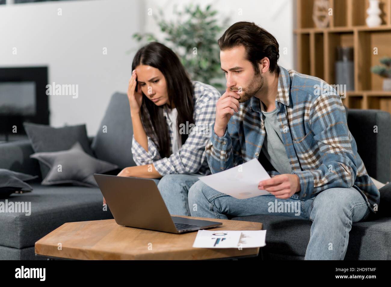 Caucasian couple sitting on the couch, the guy and the girl are freelancers, work from home, use a laptop, look at graphs, are unhappy with the results, low profit, thinking about strategy Stock Photo