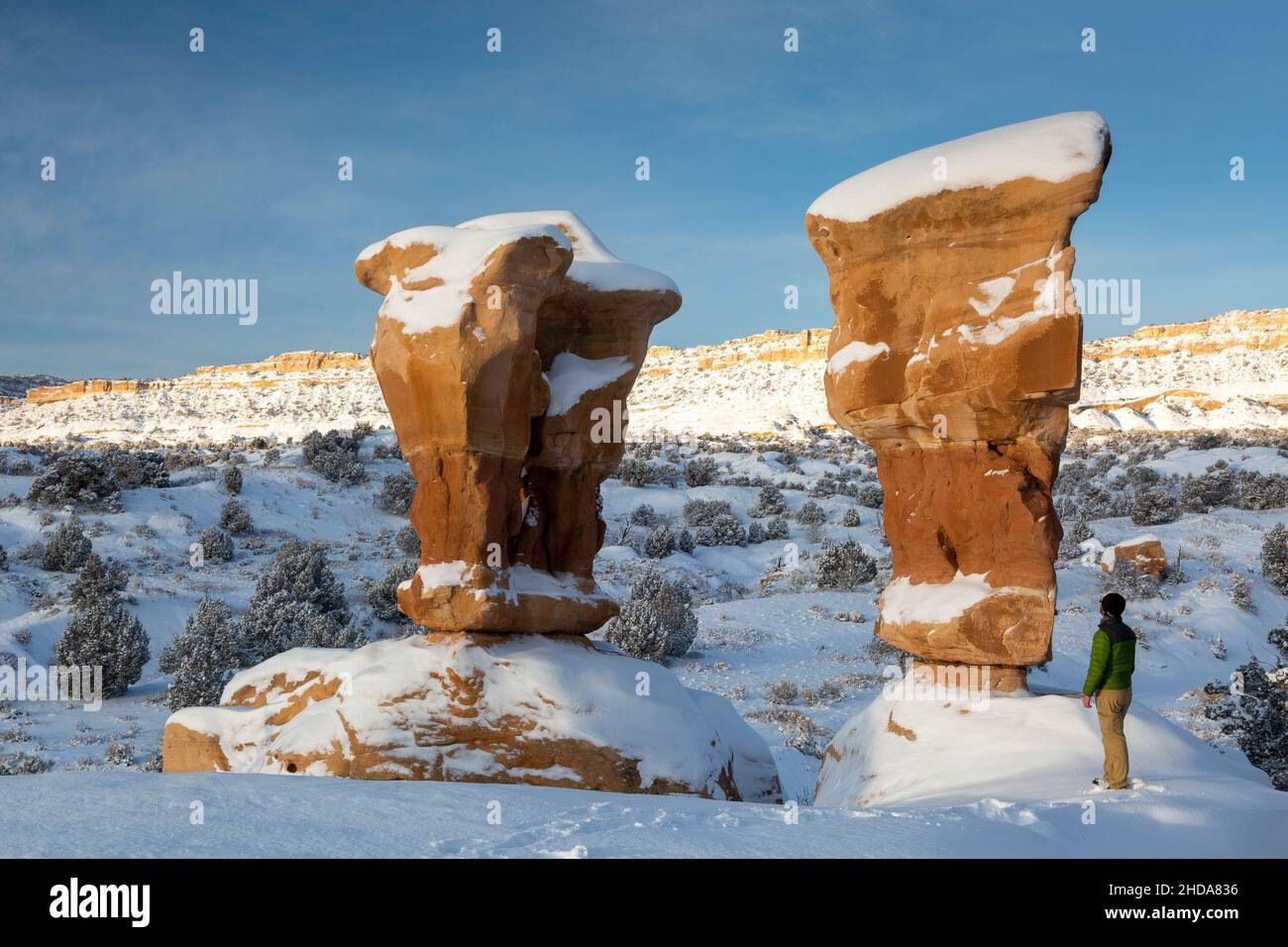 A male hiker standing below two large hoodoos covered in snow at Devils Garden. Grand Staircase-Escalante National Monument, Utah Stock Photo