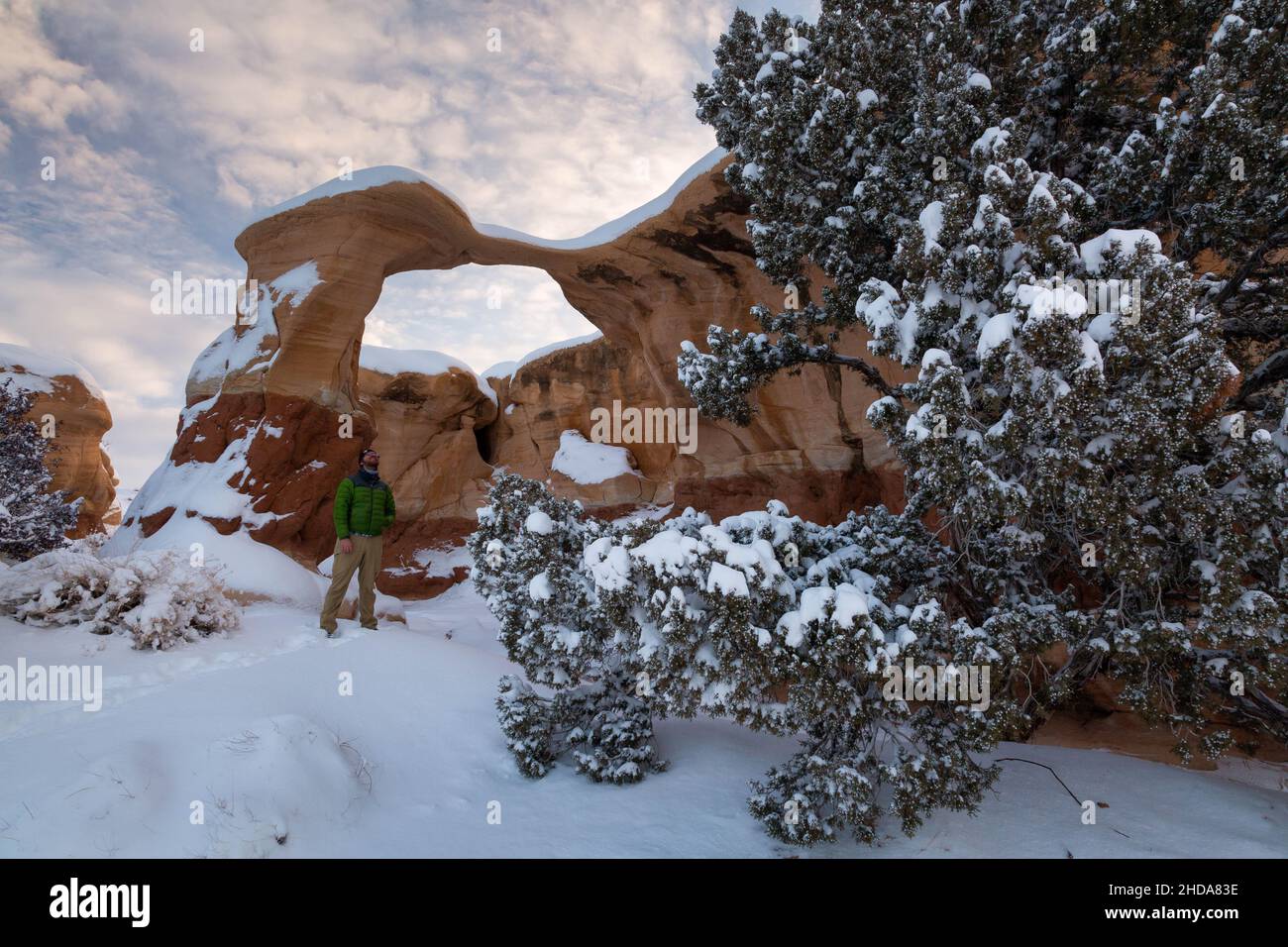 A male hiker standing below a snow-covered Metate Arch at sunrise. Grand Staircase-Escalante National Monument, Utah Stock Photo