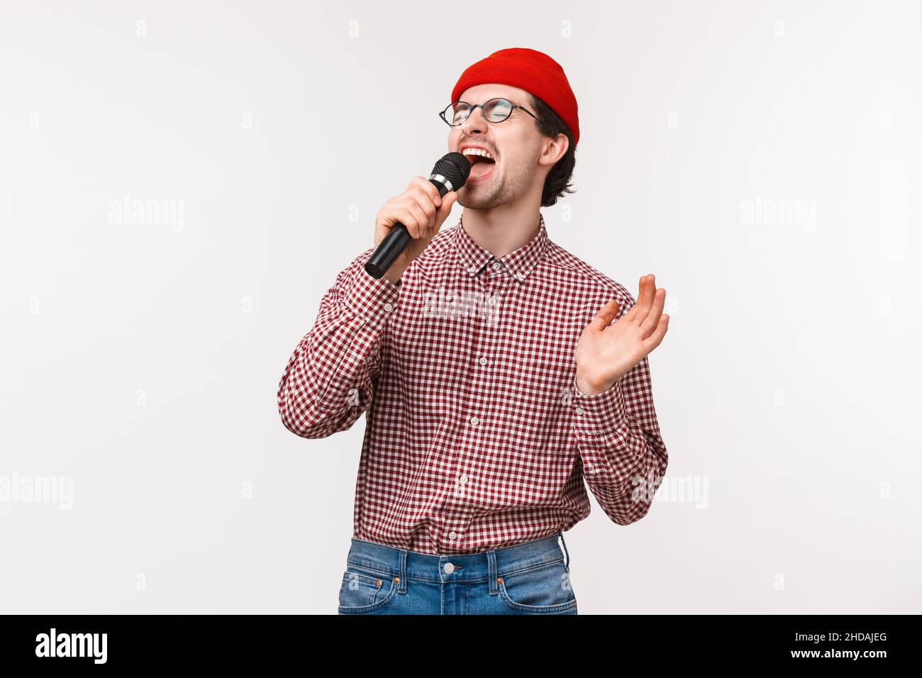 Brokenhearted guy singing romantic song in karaoke with passionate feeling, close eyes and scream into microphone, standing in red beanie, glasses and Stock Photo