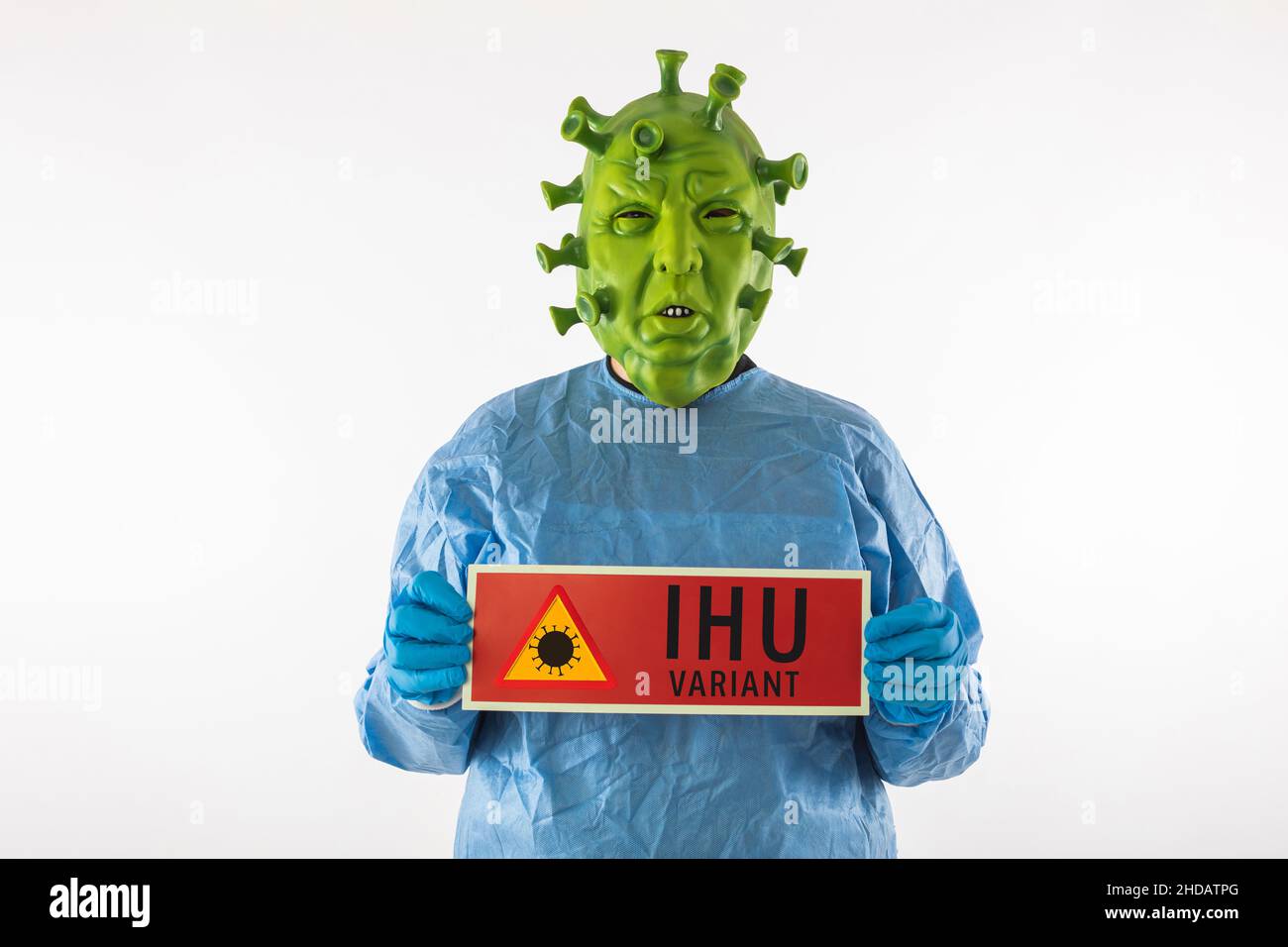 Man dressed in coronavirus latex mask, PPE and latex gloves, holding a red danger sign that reads: 'IHU variant', on white background. Coronavirus, he Stock Photo