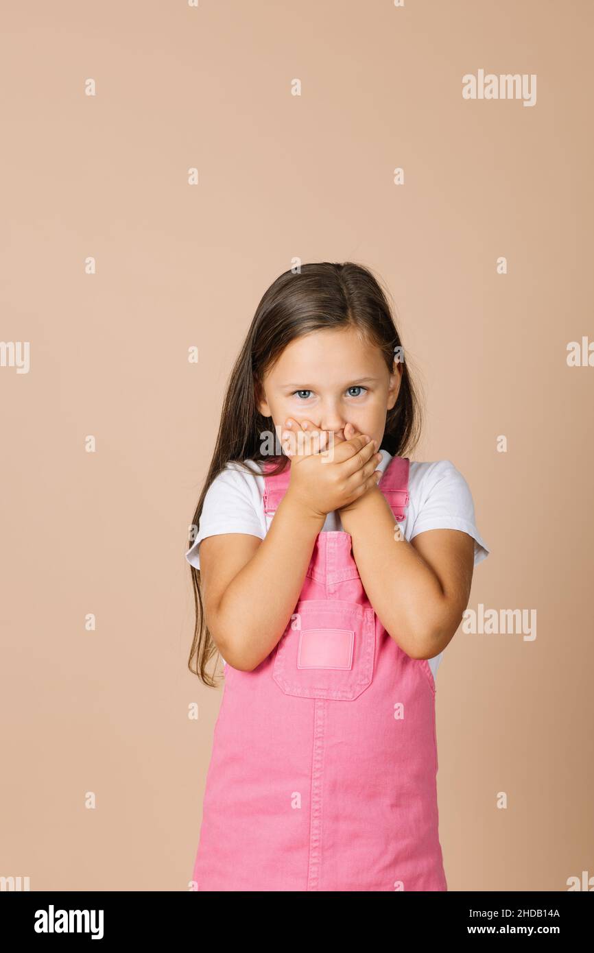 Portrait photo of female kid closing mouth with hands not talk with others with shining eyes and sullen look looking at camera wearing bright pink Stock Photo