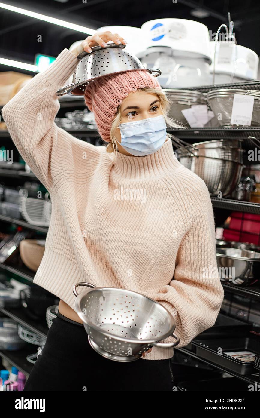 Young woman in mask, pink knitted hat and sweater is indulging in supermarket with two colanders in kitchen utensils department, wearing one on her he Stock Photo