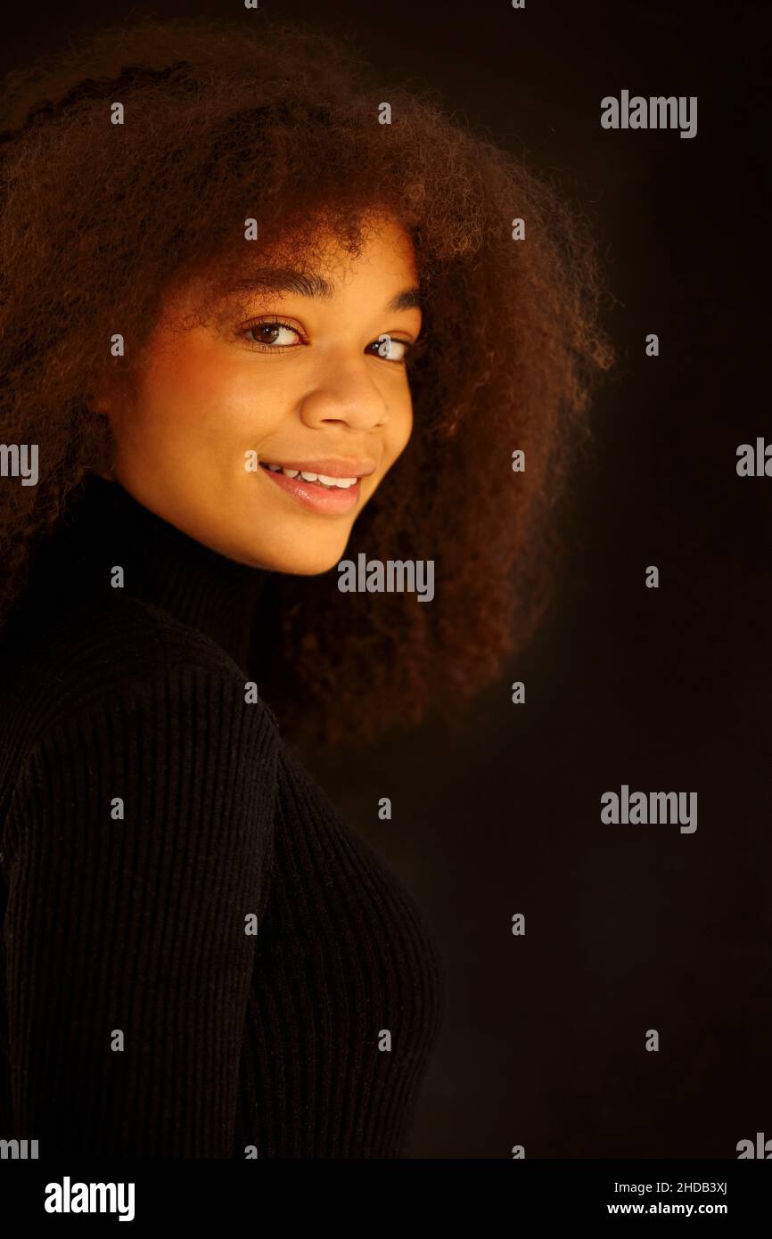 Portrait of attractive smiling curly african-american woman in black clothes looking at camera, posing isolated on dark studio background. Young natur Stock Photo