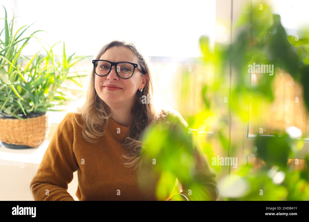 Portrait of smiling middle-aged woman wearing glasses sitting in room with green potted houseplants, selective focus. Positive happy 40s female teache Stock Photo
