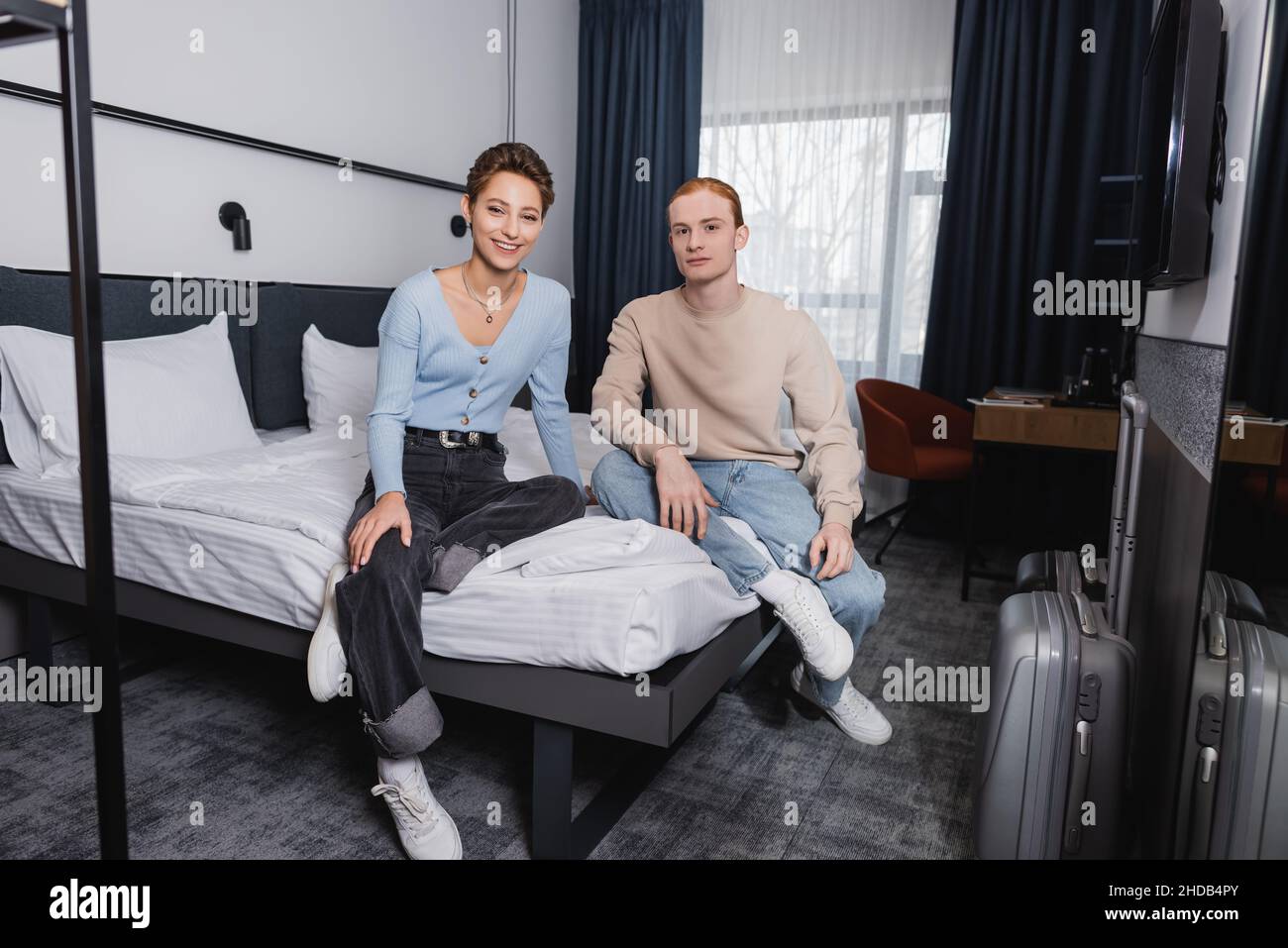 Smiling woman sitting on bed near boyfriend and suitcases in hotel room ...