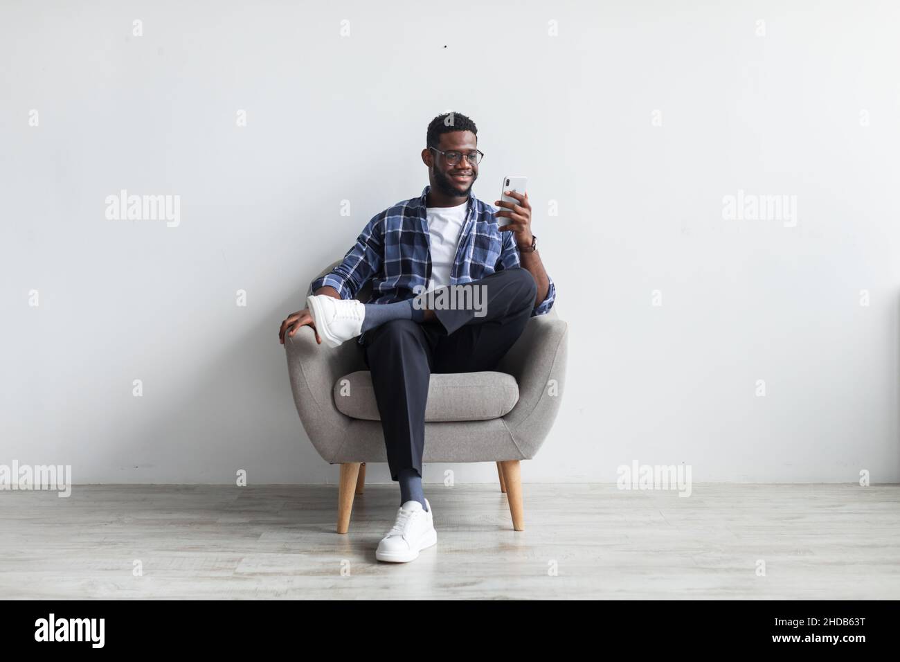 Happy black guy sitting in armchair with smartphone, texting to friend ...