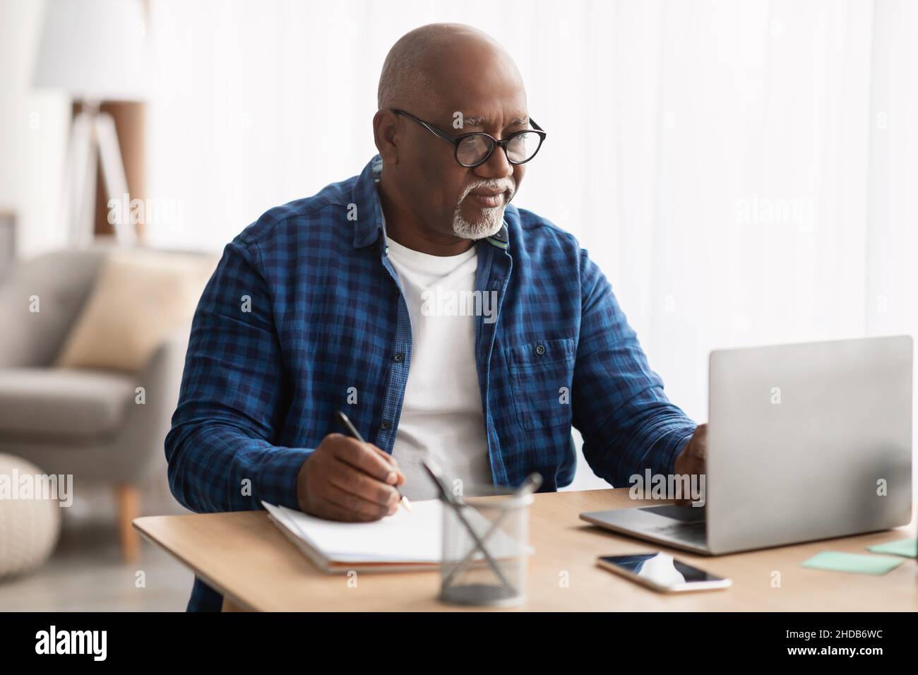 Black Man Using Laptop Taking Notes In Office, Wearing Eyeglasses Stock Photo