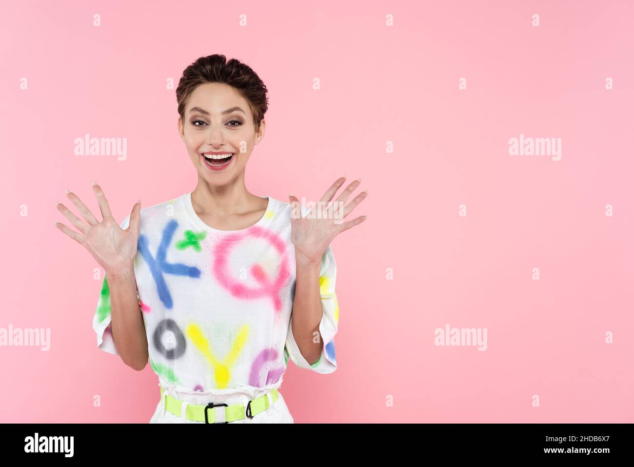 excited and trendy woman waving hands while smiling at camera isolated on pink Stock Photo