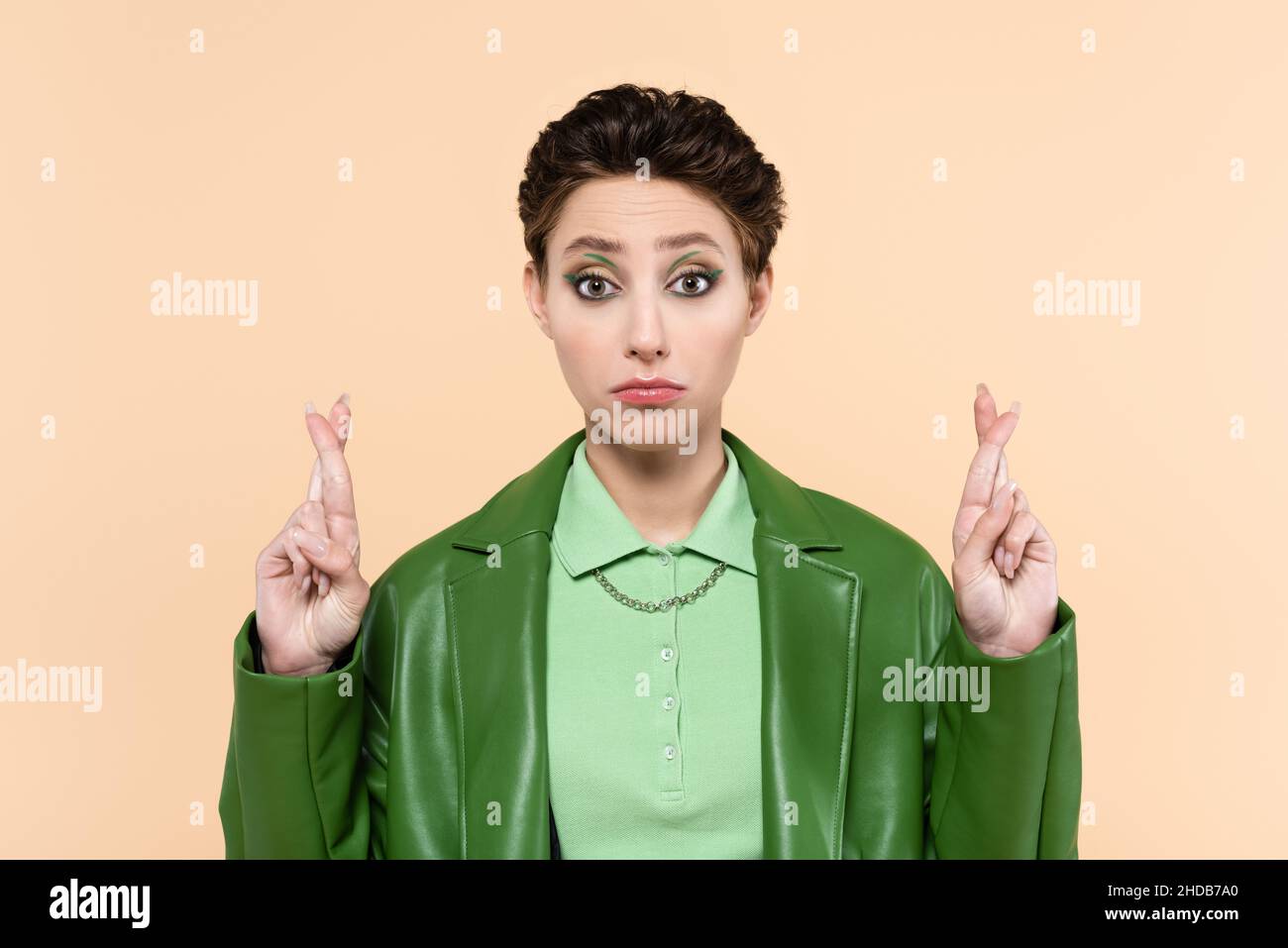 discouraged woman in green leather jacket holding crossed fingers isolated on beige Stock Photo