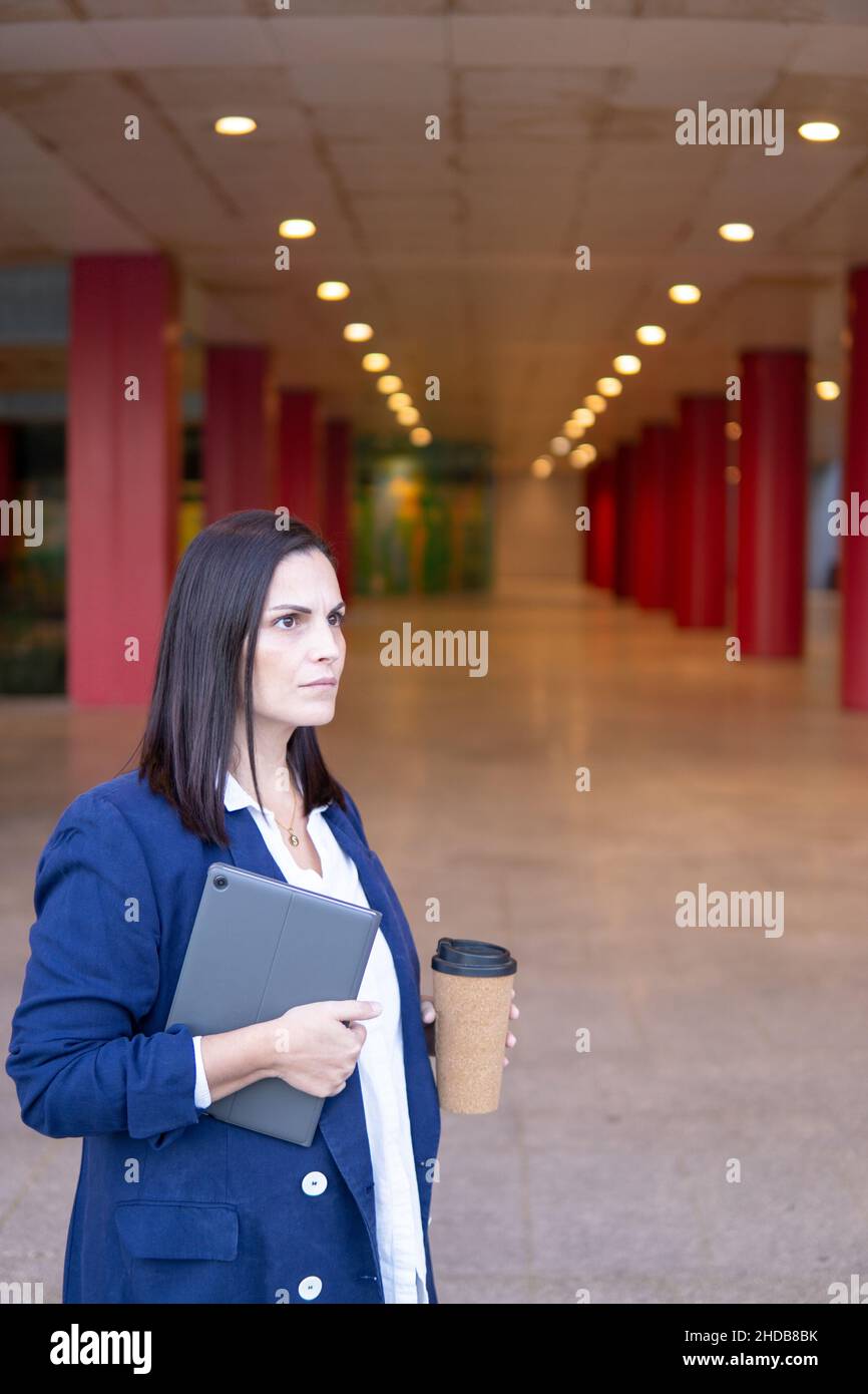 Beautiful Caucasian businesswoman with a cup of coffe and tablet working in the street outside of an office building, vertical shot Stock Photo