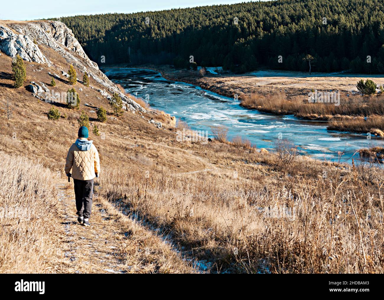 rear view of woman in neutral colors clothes walking by river in autumn on dry grass against rocks and forest, earth tones landscape, hiking enjoying Stock Photo