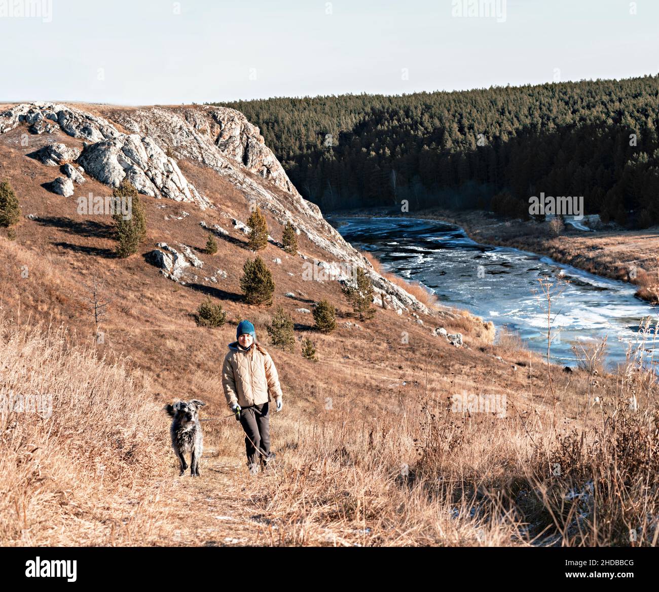 young woman in neutral colors clothes walking by river with gray fluffy mixed breed senior dog in autumn on dry grass, pet love friendship and care , Stock Photo