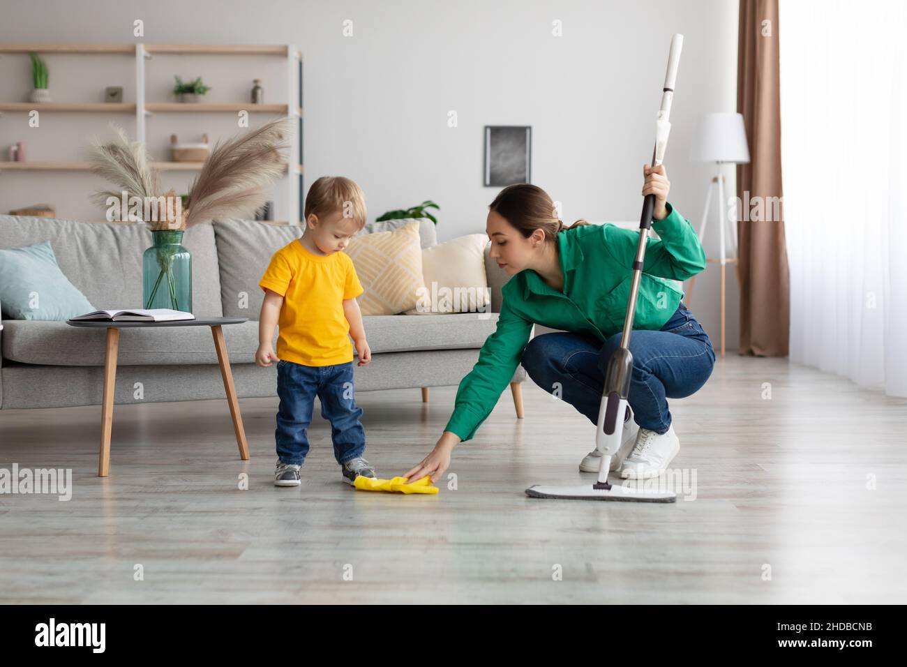 Little kid boy watching her mother mopping the floor after him. Woman wiping the floor with rag next to her baby Stock Photo