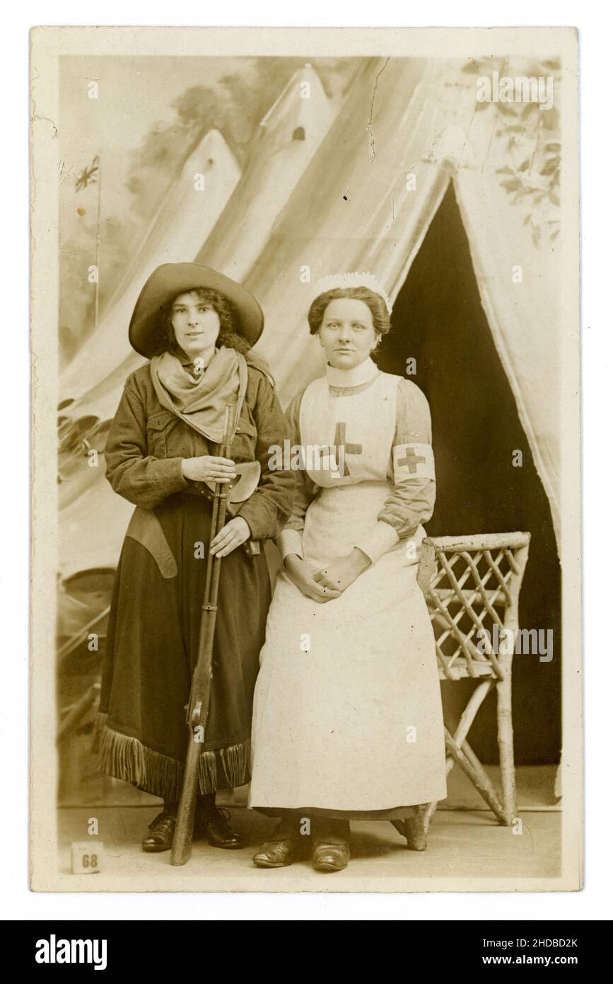 Original WW1 era postcard of 2 attractive young women, friends, having fun, posing for a photo in fancy dress costume one is dressed as cowboy / cowgirl with wide brimmed hat holding a rifle, and the other as the iconic red cross nurse - from a popular London studio called the Fancy Dress Studio at 49 Tottenham Court Road, London  circa 1914, 1915 Stock Photo